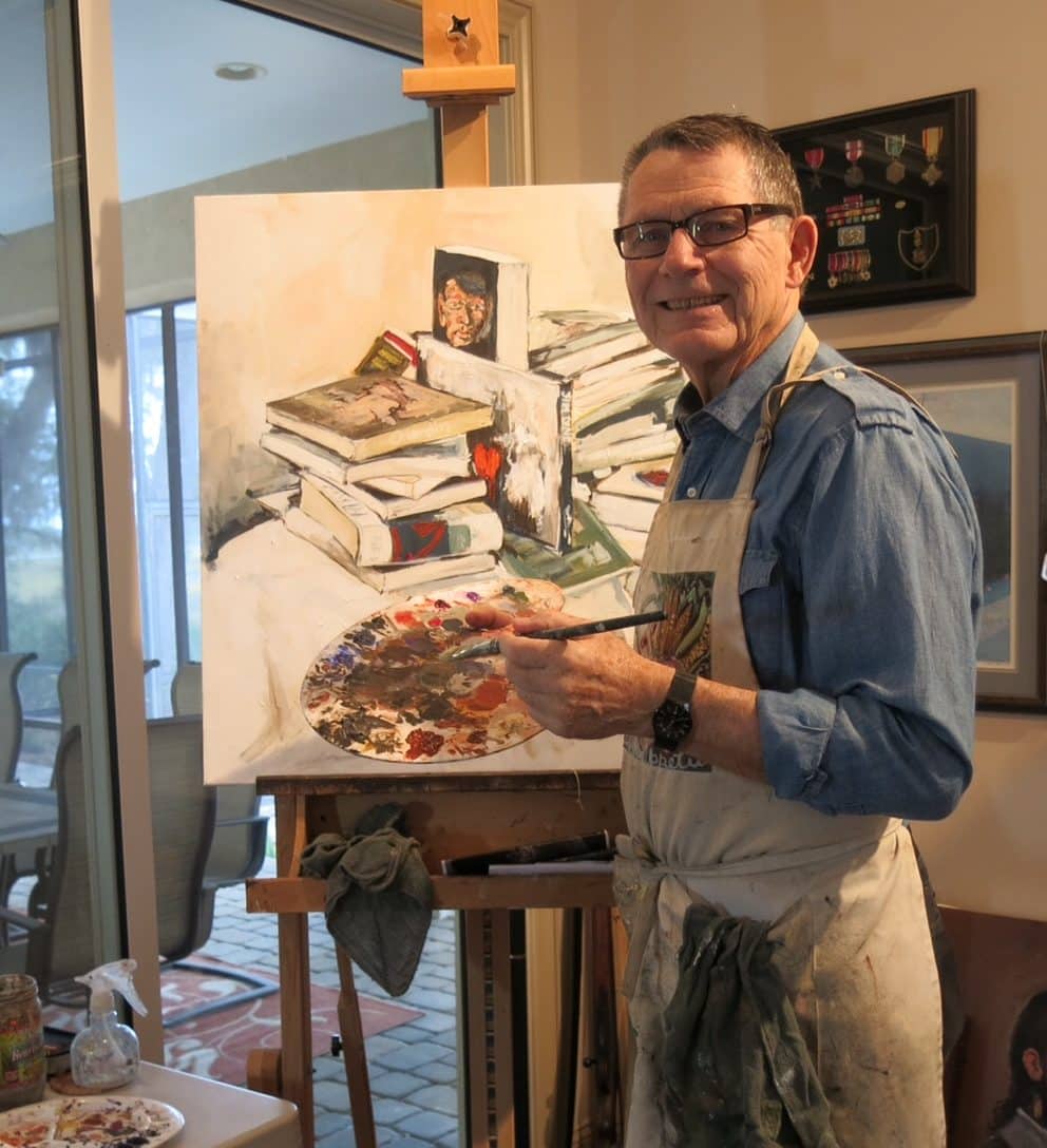 man, jerry domask, dressed in a paint covered apron holding a painting pallete standing in front of an easel with a painting of books